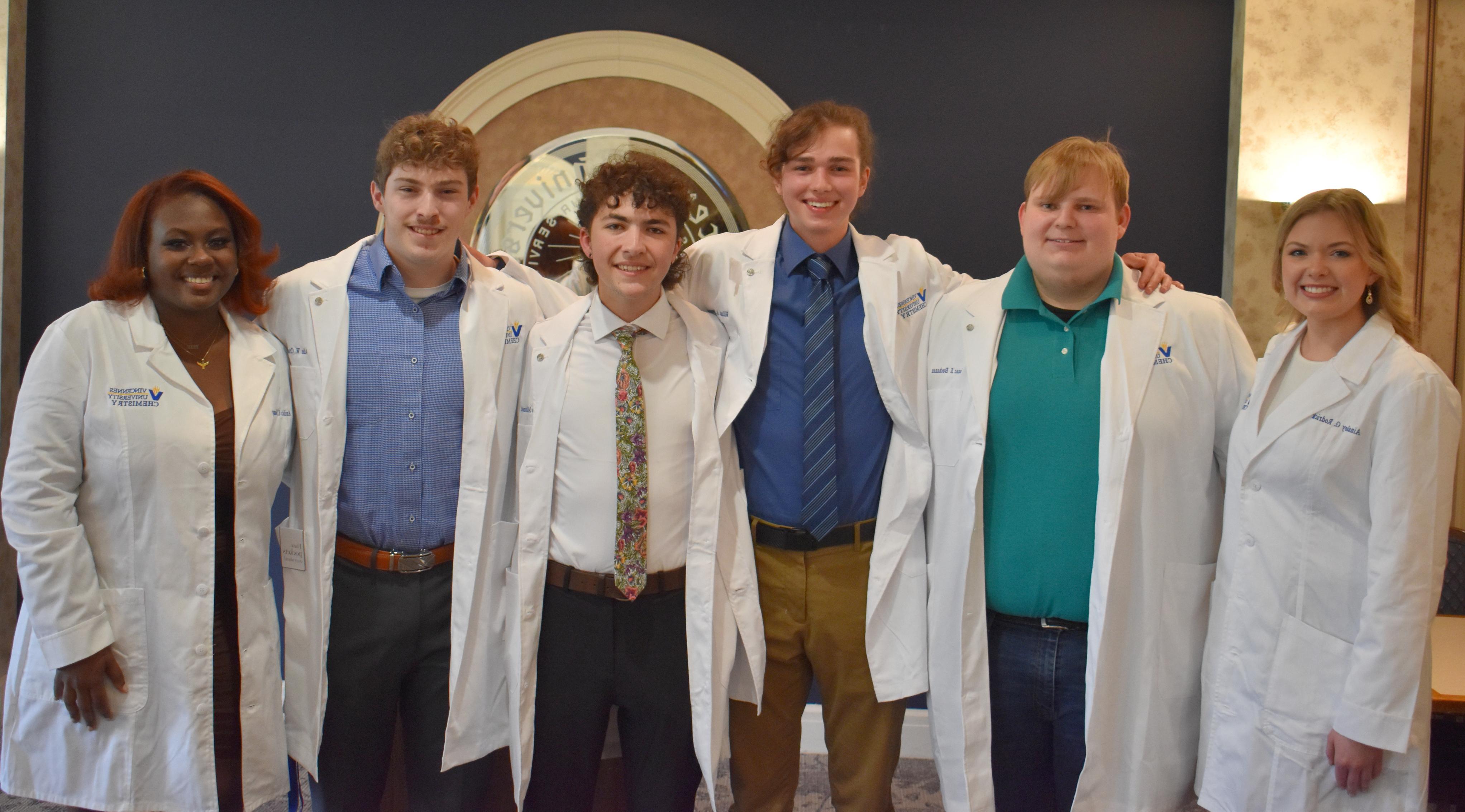 A group of six diverse male and female students wearing white labs with their arms wrapped around each other pose for a photo.